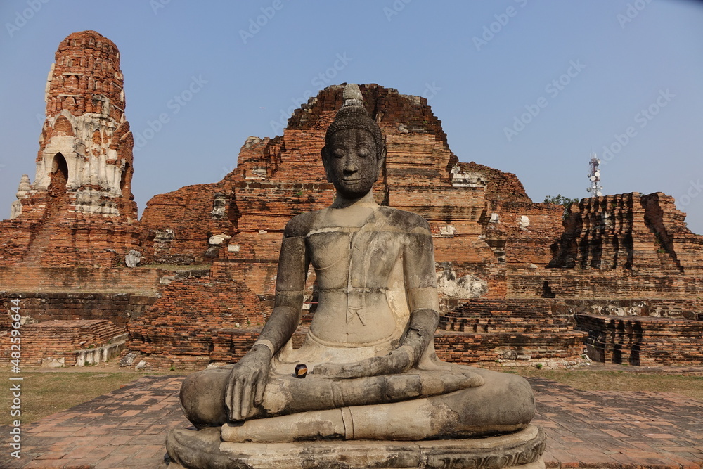 Buddha statue at Wat Mahathat with small black pot resting on its leg and the ruins in the background (horizontal image), Ayutthaya, Thailand Buddha statue at Wat Mahathat with small black pot resting on its leg and the ruins in the background (horizontal image), Ayutthaya, Thailand