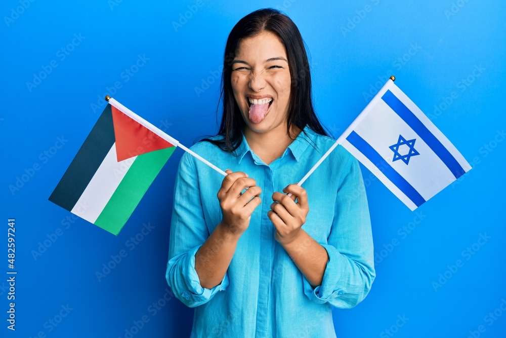 Young latin woman holding palestine and israel flags sticking tongue out happy with funny expression. Young latin woman holding palestine and israel flags sticking tongue out happy with funny expression.