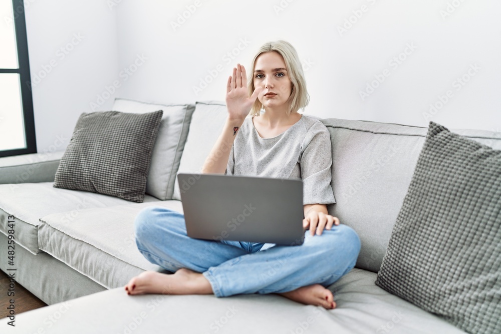 Young caucasian woman using laptop at home sitting on the sofa doing stop sing with palm of the hand. warning expression with negative and serious gesture on the face.