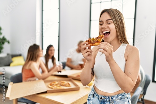 Group of young people eating italian pizza sitting on the table at home. Woman standing and holding portion.