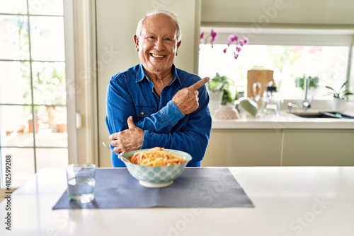 Senior man with grey hair eating pasta spaghetti at home with a big smile on face, pointing with hand and finger to the side looking at the camera.