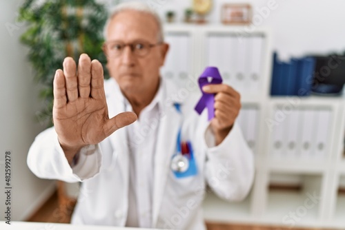 Senior doctor man holding purple ribbon awareness with open hand doing stop sign with serious and confident expression, defense gesture