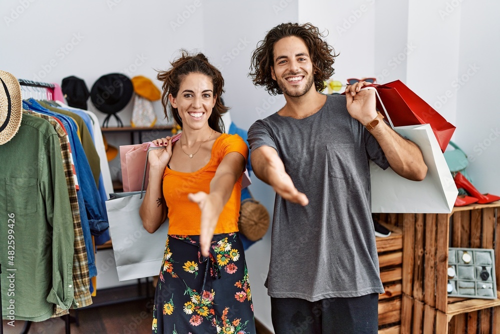 Obraz premium Young hispanic couple holding shopping bags at retail shop smiling friendly offering handshake as greeting and welcoming. successful business.