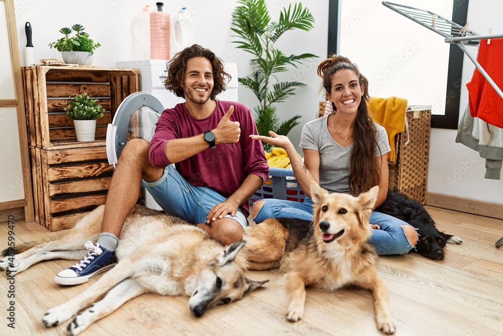 Young hispanic couple doing laundry with dogs showing palm hand and doing ok gesture with thumbs up, smiling happy and cheerful