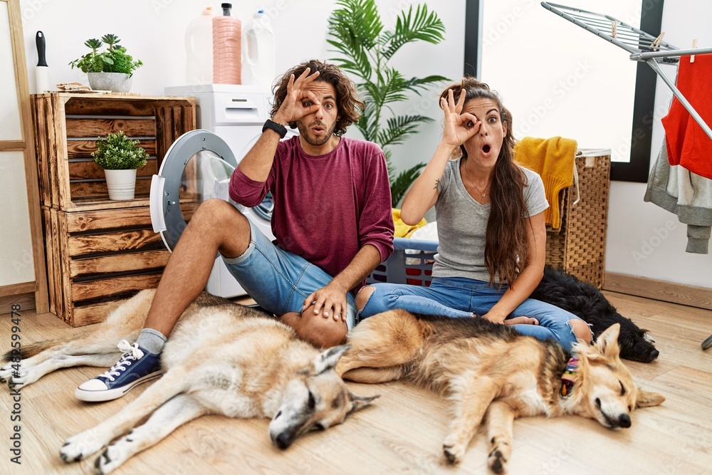 Young hispanic couple doing laundry with dogs doing ok gesture shocked with surprised face, eye looking through fingers. unbelieving expression.