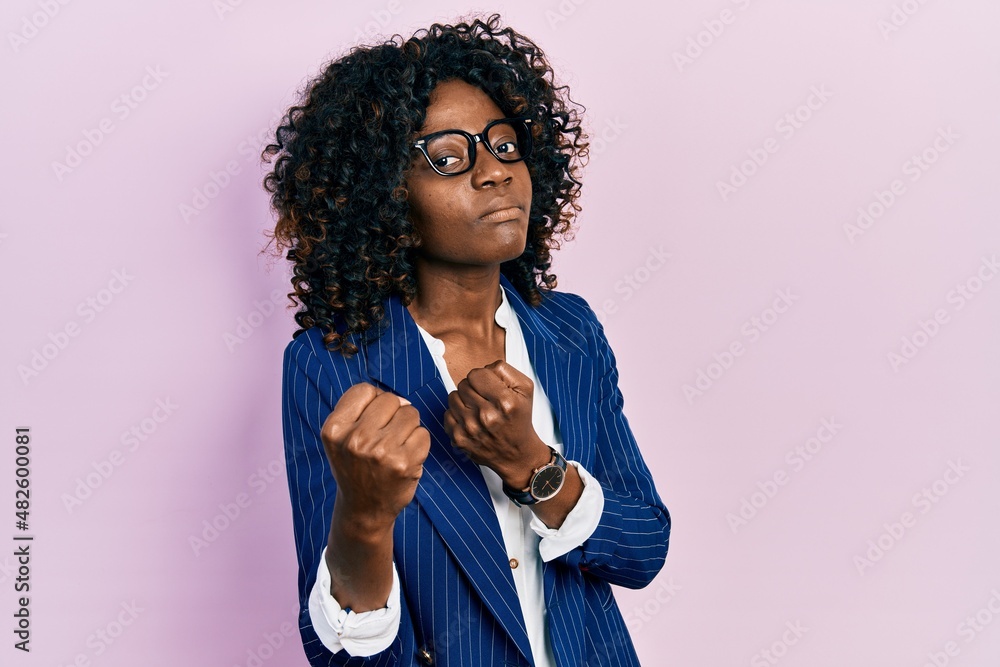 Young african american woman wearing business clothes and glasses ready to fight with fist defense gesture, angry and upset face, afraid of problem Young african american woman wearing business clothes and glasses ready to fight with fist defense gesture, angry and upset face, afraid of problem