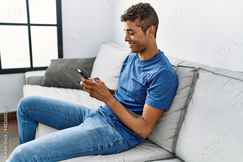 Young hispanic man relaxing sitting on the sofa using smartphone at home