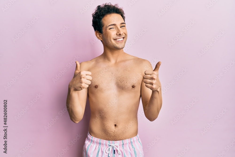 Fototapeta premium Young handsome man wearing swimwear shirtless success sign doing positive gesture with hand, thumbs up smiling and happy. cheerful expression and winner gesture.