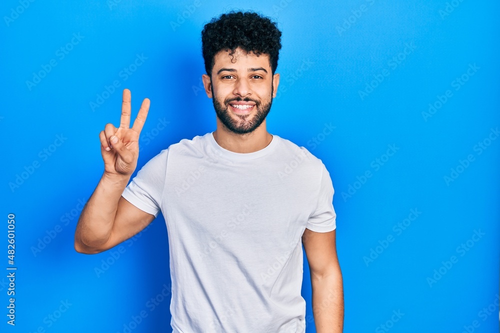 Naklejka premium Young arab man with beard wearing casual white t shirt smiling looking to the camera showing fingers doing victory sign. number two.