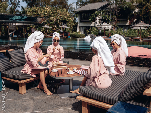 The four BFFs sitting at the table and enjoying the morning in robes, towels and sunglasses next to the pool and villas while holding cups of coffee in their hands. Holiday concept photo