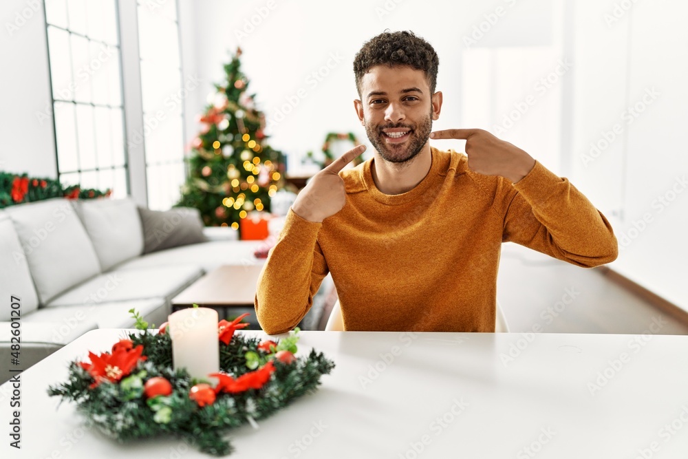 Arab young man sitting on the table by christmas tree smiling cheerful showing and pointing with fingers teeth and mouth. dental health concept. Arab young man sitting on the table by christmas tree smiling cheerful showing and pointing with fingers teeth and mouth. dental health concept.