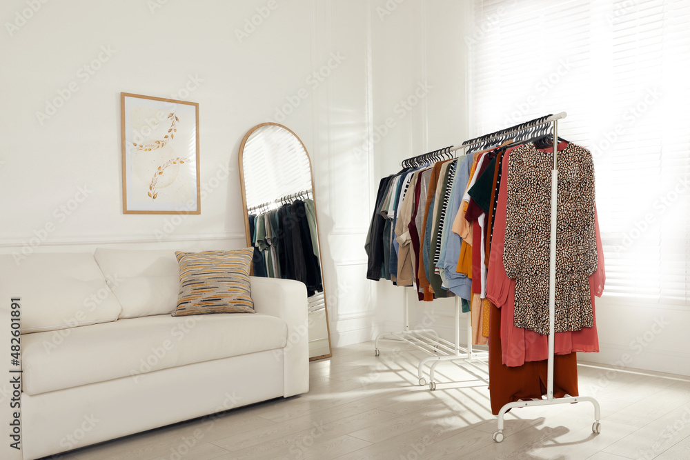 Rack with stylish clothes near sofa and mirror indoors. Fast fashion