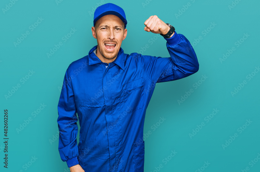 Bald man with beard wearing builder jumpsuit uniform angry and mad raising fist frustrated and furious while shouting with anger. rage and aggressive concept.