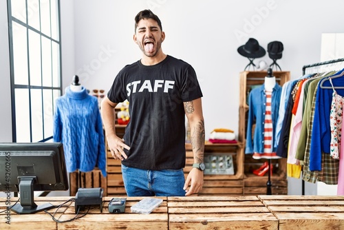 Young hispanic man working at retail boutique sticking tongue out happy with funny expression. emotion concept.