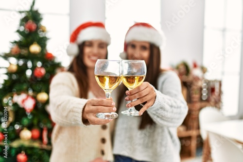 Two women toasting with champagne and hugging each other standing by christmas tree at home