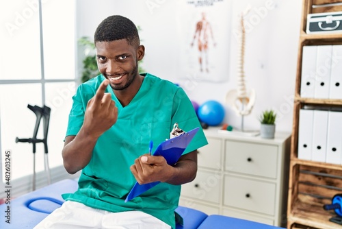 Young african american man working at pain recovery clinic beckoning come here gesture with hand inviting welcoming happy and smiling