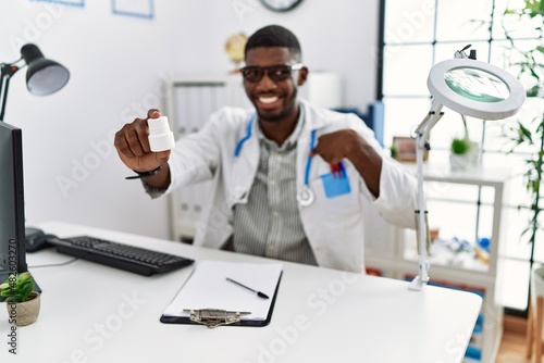 Young african american doctor man wearing doctor uniform holding prescription pills at the clinic pointing finger to one self smiling happy and proud