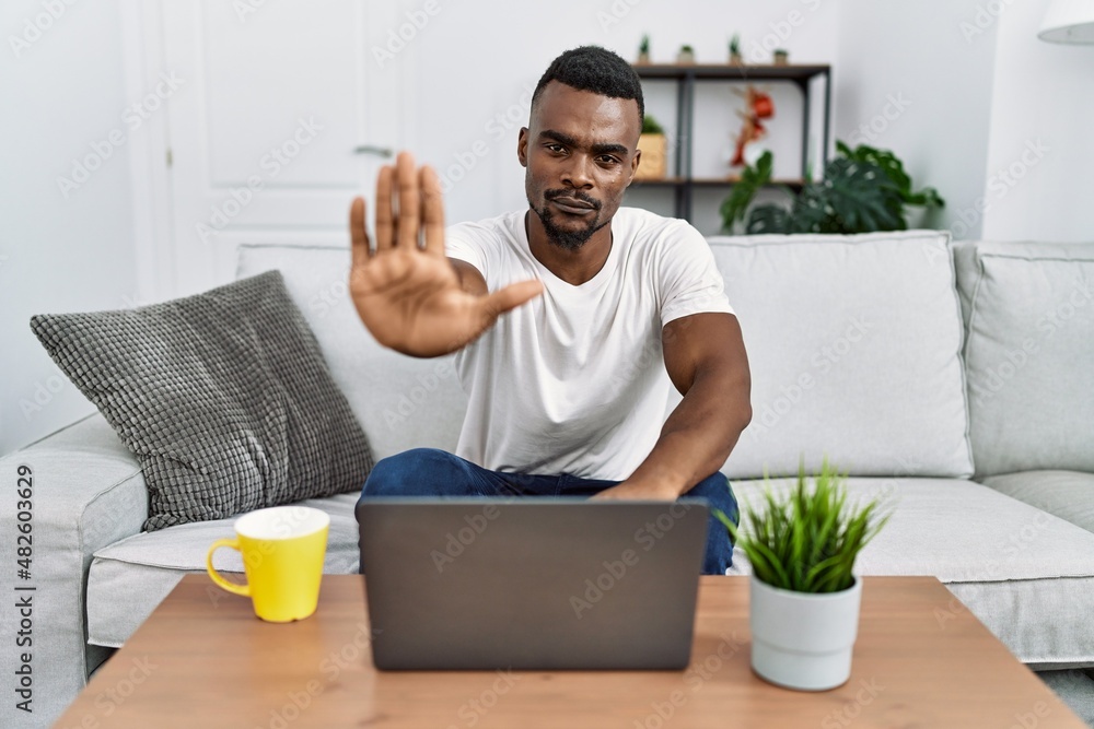 Young african man using laptop at home doing stop sing with palm of the hand. warning expression with negative and serious gesture on the face. Young african man using laptop at home doing stop sing with palm of the hand. warning expression with negative and serious gesture on the face.