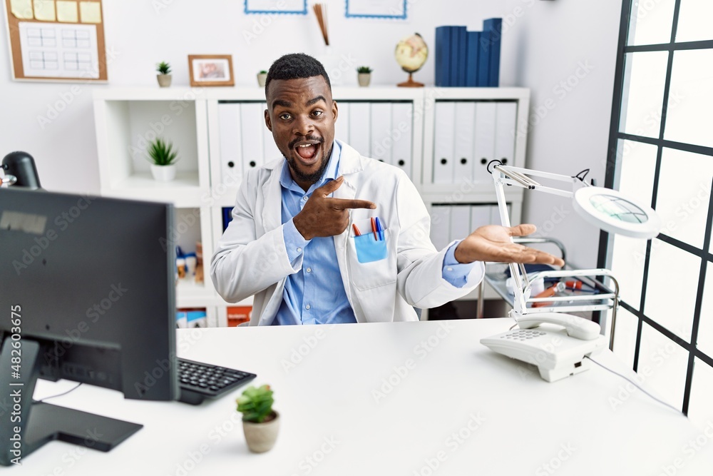 Young african doctor man wearing stethoscope at the clinic amazed and smiling to the camera while presenting with hand and pointing with finger.