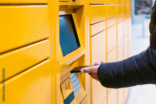 Detail of the arm of a man scanning a code on the mobile phone to pick up a package from the yellow locker. Messaging concept, compare online, e-commerce and packages