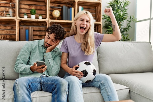 Woman smiling happy watching soccer match and boyfriend boring using smartphone at home.