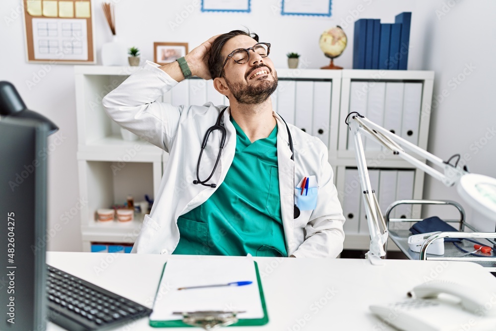 Young man with beard wearing doctor uniform and stethoscope at the clinic smiling confident touching hair with hand up gesture, posing attractive and fashionable Young man with beard wearing doctor uniform and stethoscope at the clinic smiling confident touching hair with hand up gesture, posing attractive and fashionable