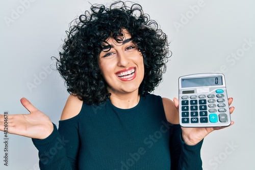 Young middle east woman showing calculator device celebrating achievement with happy smile and winner expression with raised hand