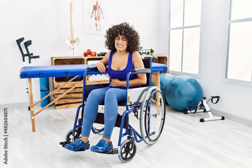 Young middle eastern woman sitting on wheelchair at physiotherapy clinic with hand on stomach because nausea, painful disease feeling unwell. ache concept. Young middle eastern woman sitting on wheelchair at physiotherapy clinic with hand on stomach because nausea, painful disease feeling unwell. ache concept.