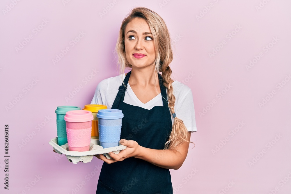 Beautiful young blonde woman wearing waitress apron holding take away cup of coffees smiling looking to the side and staring away thinking. Beautiful young blonde woman wearing waitress apron holding take away cup of coffees smiling looking to the side and staring away thinking.