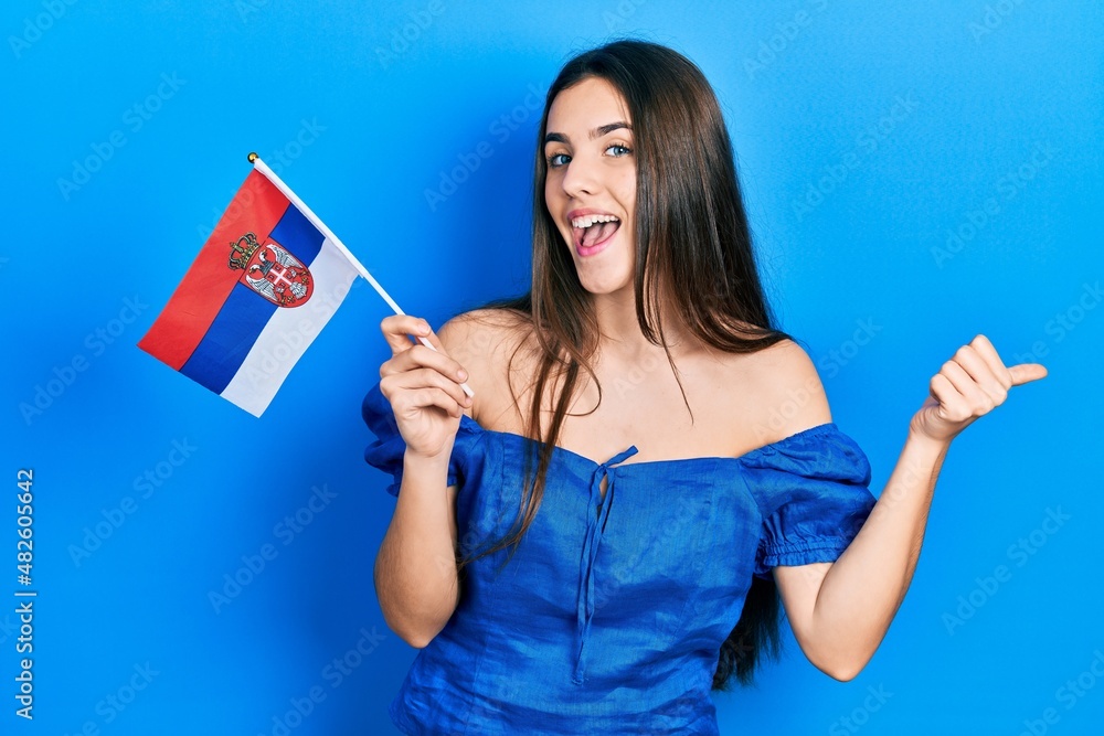 Young brunette teenager holding serbia flag pointing thumb up to the side smiling happy with open mouth Young brunette teenager holding serbia flag pointing thumb up to the side smiling happy with open mouth