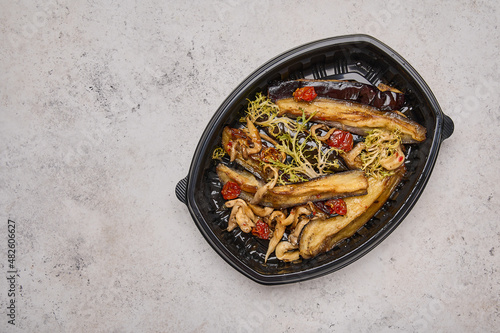 Crispy eggplant with baked cherry tomatoes and oyster mushrooms in black plastic container on light background top view. Designer food concept for take away or delivery