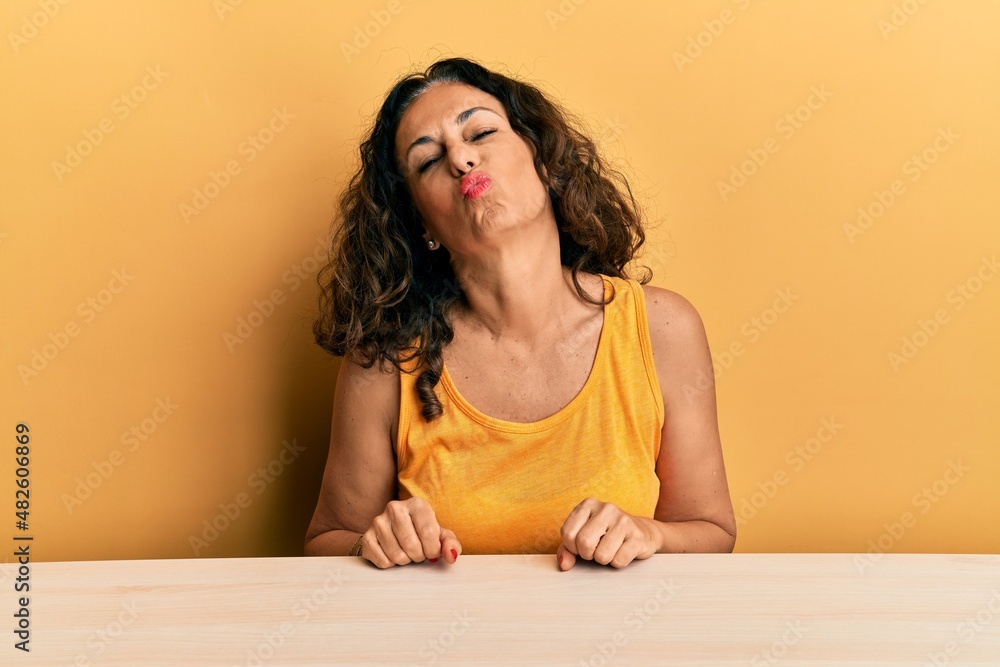 Beautiful middle age woman wearing casual clothes sitting on the table looking at the camera blowing a kiss on air being lovely and sexy. love expression. Beautiful middle age woman wearing casual clothes sitting on the table looking at the camera blowing a kiss on air being lovely and sexy. love expression.