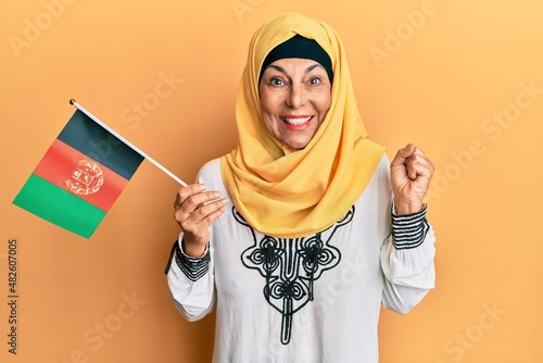 Middle age hispanic woman holding afghanistan flag screaming proud, celebrating victory and success very excited with raised arm