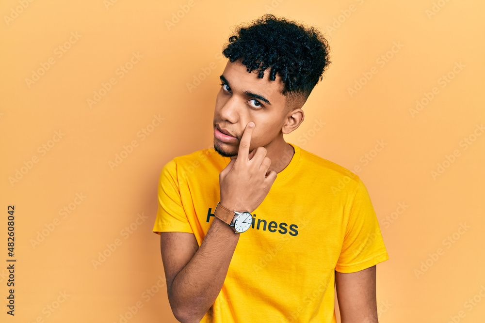 Young african american man wearing t shirt with happiness word message pointing to the eye watching you gesture, suspicious expression Young african american man wearing t shirt with happiness word message pointing to the eye watching you gesture, suspicious expression