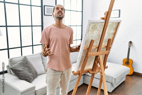 Young hispanic man with beard painting on canvas at home angry and mad screaming frustrated and furious, shouting with anger. rage and aggressive concept.