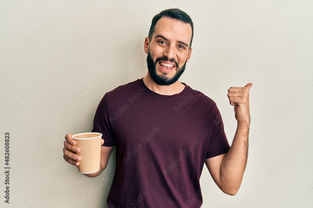 Young man with beard drinking a take away cup coffee pointing thumb up to the side smiling happy with open mouth Young man with beard drinking a take away cup coffee pointing thumb up to the side smiling happy with open mouth