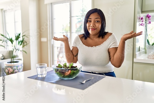 Young hispanic woman eating healthy salad at home clueless and confused expression with arms and hands raised. doubt concept.