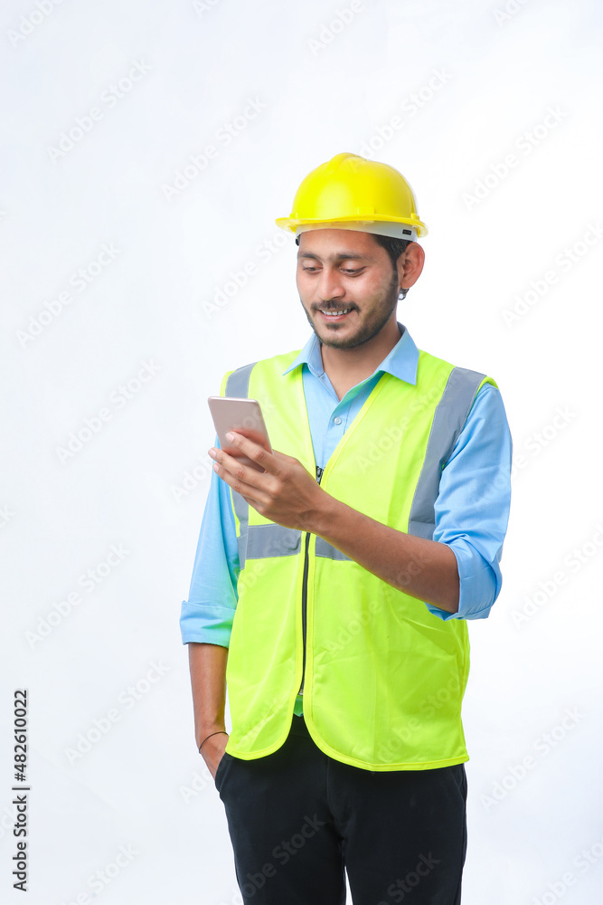 Young indian engineer wearing hardhat and using smartphone on white background.