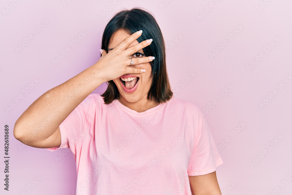 Young hispanic woman wearing casual pink t shirt peeking in shock covering face and eyes with hand, looking through fingers with embarrassed expression. Young hispanic woman wearing casual pink t shirt peeking in shock covering face and eyes with hand, looking through fingers with embarrassed expression.