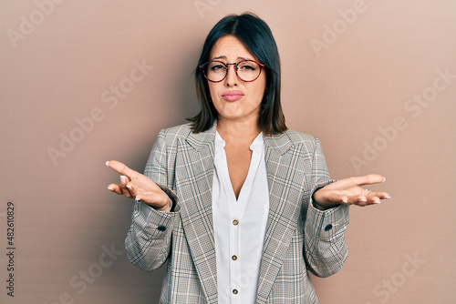 Young hispanic woman wearing business clothes and glasses clueless and confused expression with arms and hands raised. doubt concept.