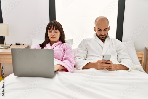 Young hispanic couple using laptop and smartphone sitting on the bed at bedroom.