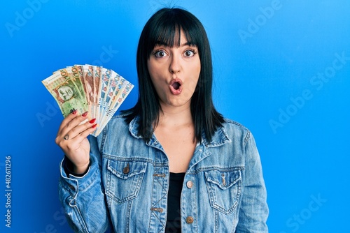 Young hispanic woman holding peruvian sol banknotes scared and amazed with open mouth for surprise, disbelief face