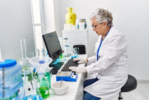 Senior grey-haired woman wearing scientist uniform using computer working at laboratory