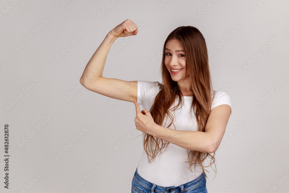 Portrait of confident independent woman raising hand pointing biceps, feeling power and energy to achieve goal, wearing white T-shirt. Indoor studio shot isolated on gray background. Portrait of confident independent woman raising hand pointing biceps, feeling power and energy to achieve goal, wearing white T-shirt. Indoor studio shot isolated on gray background.