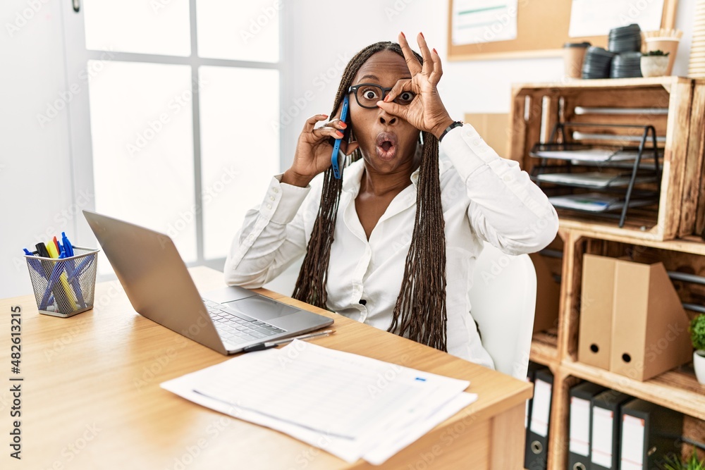 Black woman with braids working at the office speaking on the phone doing ok gesture shocked with surprised face, eye looking through fingers. unbelieving expression.