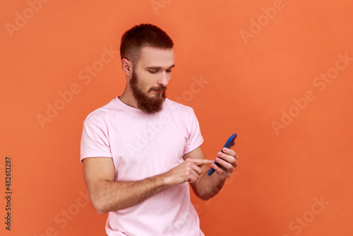 Portrait of bearded man scrolling online, texting message in social media on cell phone, using mobile network services, wearing pink T-shirt. Indoor studio shot isolated on orange background. © khosrork