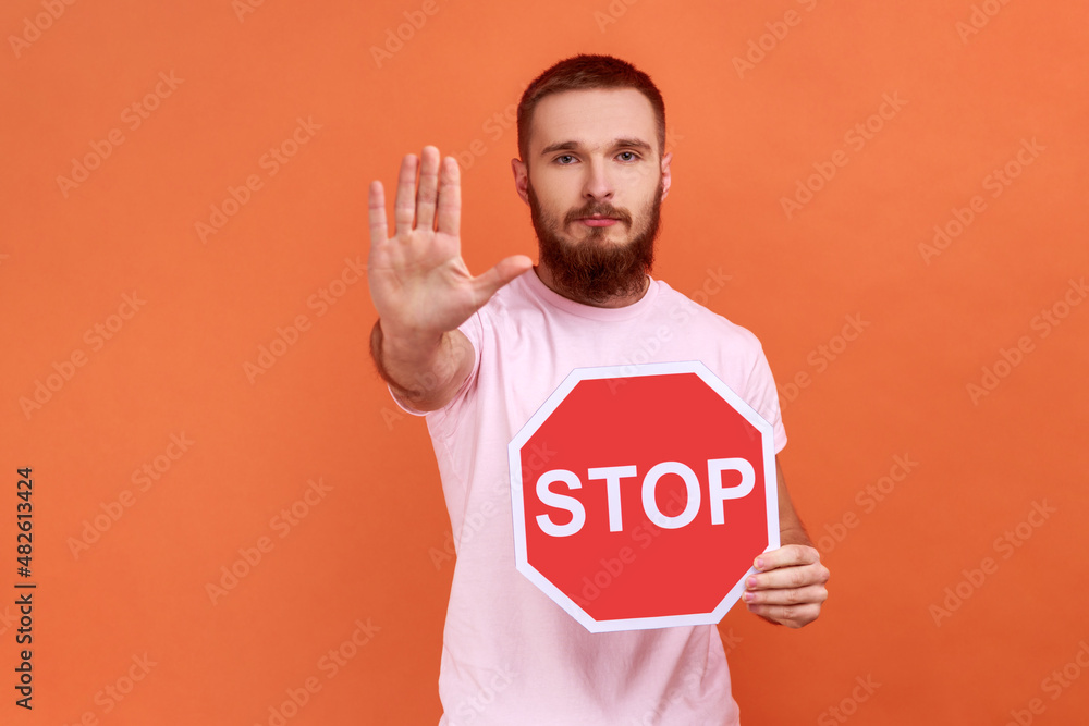 Portrait of strict bossy bearded man showing stop gesture and holding red stop road sign, prohibitions and restrictions, wearing pink T-shirt. Indoor studio shot isolated on orange background. Portrait of strict bossy bearded man showing stop gesture and holding red stop road sign, prohibitions and restrictions, wearing pink T-shirt. Indoor studio shot isolated on orange background.