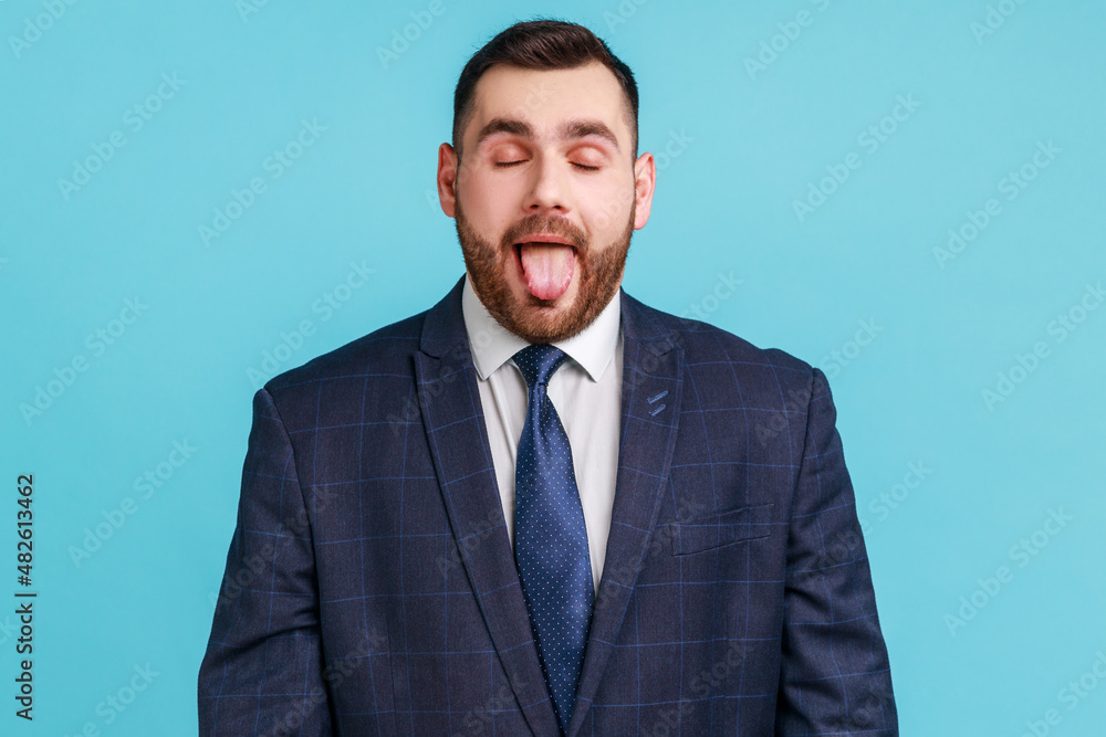 Portrait of childish carefree brunette man wearing official style suit showing out tongue and closing eyes with naughty disobedient grimace, making face. Indoor studio shot isolated on blue background Portrait of childish carefree brunette man wearing official style suit showing out tongue and closing eyes with naughty disobedient grimace, making face. Indoor studio shot isolated on blue background