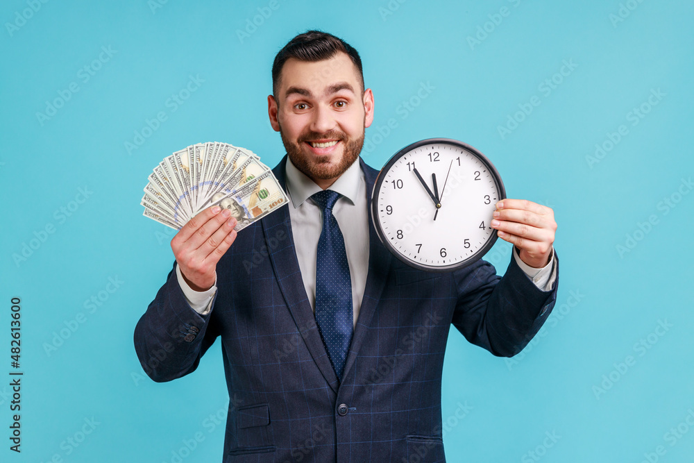 Portrait of happy businessman wearing official style suit showing dollar cash and wall clock on camera, hourly payment, looking at camera. Indoor studio shot isolated on blue background. Portrait of happy businessman wearing official style suit showing dollar cash and wall clock on camera, hourly payment, looking at camera. Indoor studio shot isolated on blue background.
