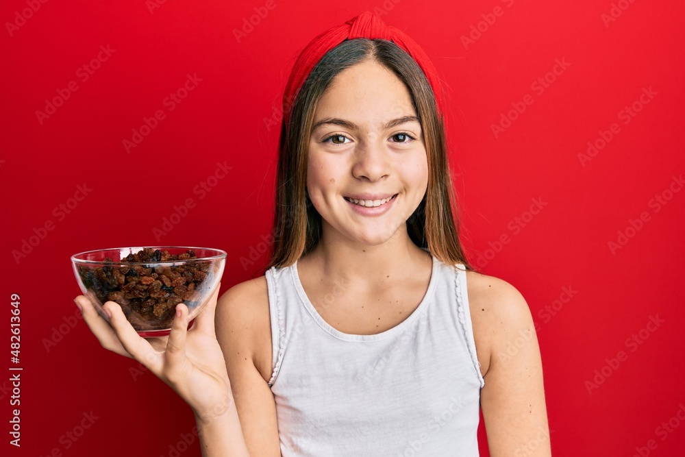 Beautiful brunette little girl holding bowl of raisins looking positive and happy standing and smiling with a confident smile showing teeth Beautiful brunette little girl holding bowl of raisins looking positive and happy standing and smiling with a confident smile showing teeth